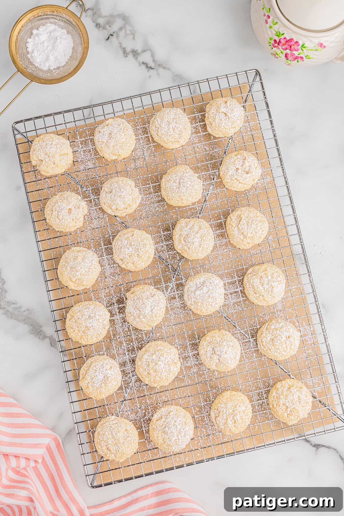 Velvety Cream Cheese Bites 6 A cooling rack filled with freshly baked cream cheese cookies, lightly sprinkled with powdered sugar, on a marble countertop with sifter and teapot.