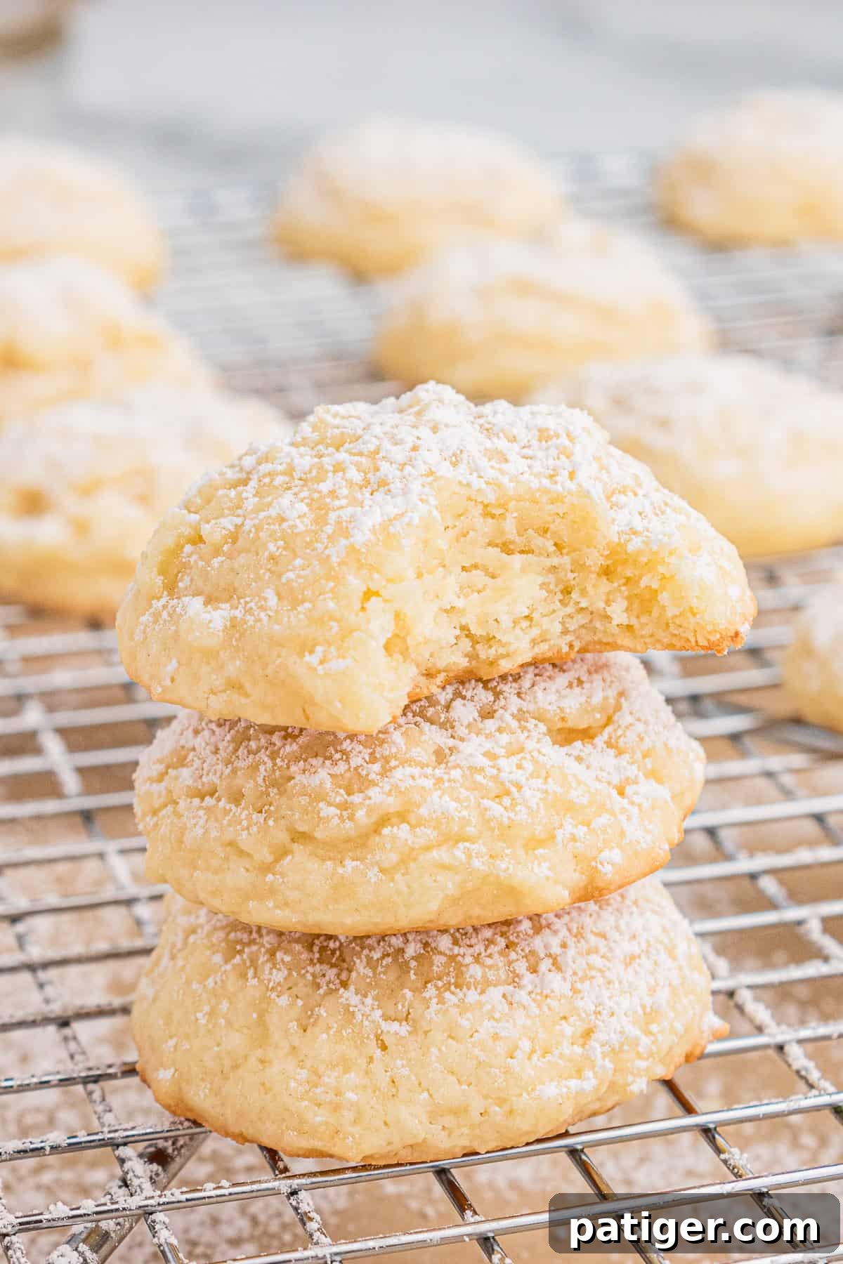 Velvety Cream Cheese Bites 7 A stack of three cream cheese cookies on a cooling rack, with a bite taken out of the top cookie to reveal the soft interior.