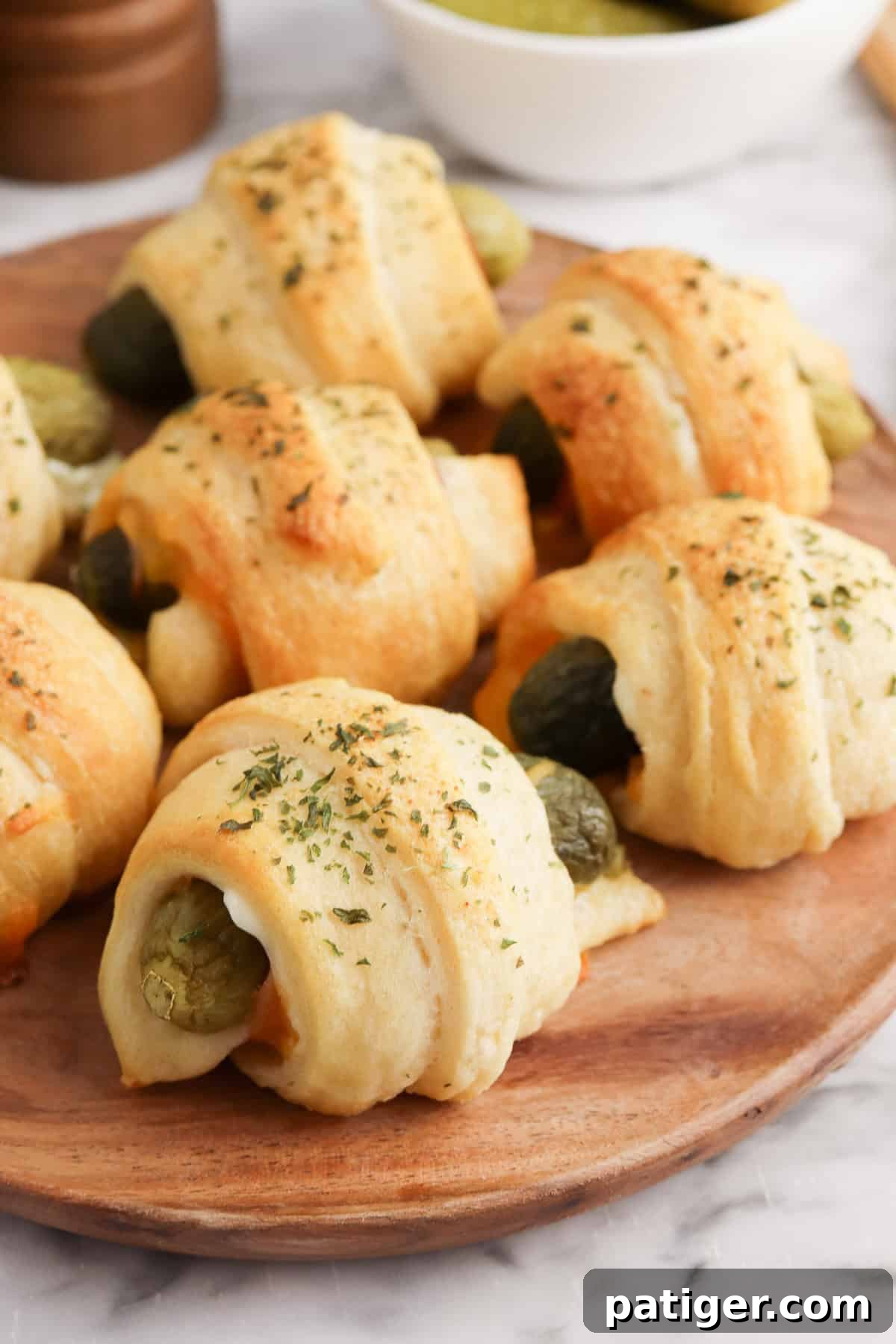 Dill Pickle Puffs 2 Golden-brown crescent roll-wrapped pickles sprinkled with dried herbs are arranged on a round wooden plate. Melted cheese is visible inside some of the rolls.