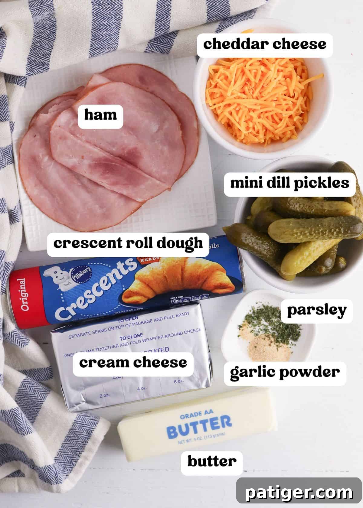 Dill Pickle Puffs 4 A flat lay of ingredients for pickles in a blanket with text labels. Visible items include slices of ham on a white plate, shredded cheddar cheese in a white bowl, a can of crescent roll dough, a block of cream cheese, mini dill pickles in a bowl, butter, and a small dish with parsley and garlic powder