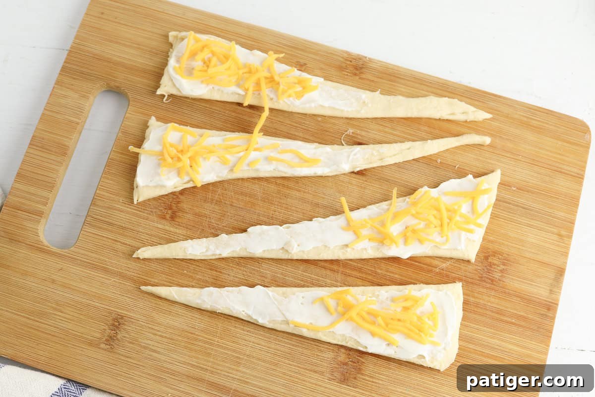 Dill Pickle Puffs 5 Four triangular pieces of crescent roll dough are laid out on a wooden cutting board. Each triangle is evenly spread with cream cheese and topped with scattered shredded cheddar cheese.