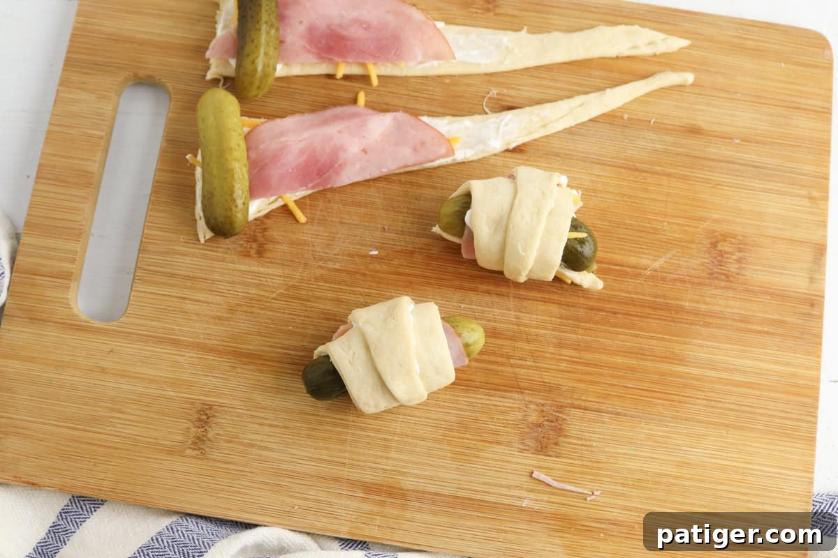 Dill Pickle Puffs 7 Two completed pickles wrapped in crescent roll dough next to two unwrapped triangles of dough layered with cream cheese, shredded cheddar, ham, and pickles.