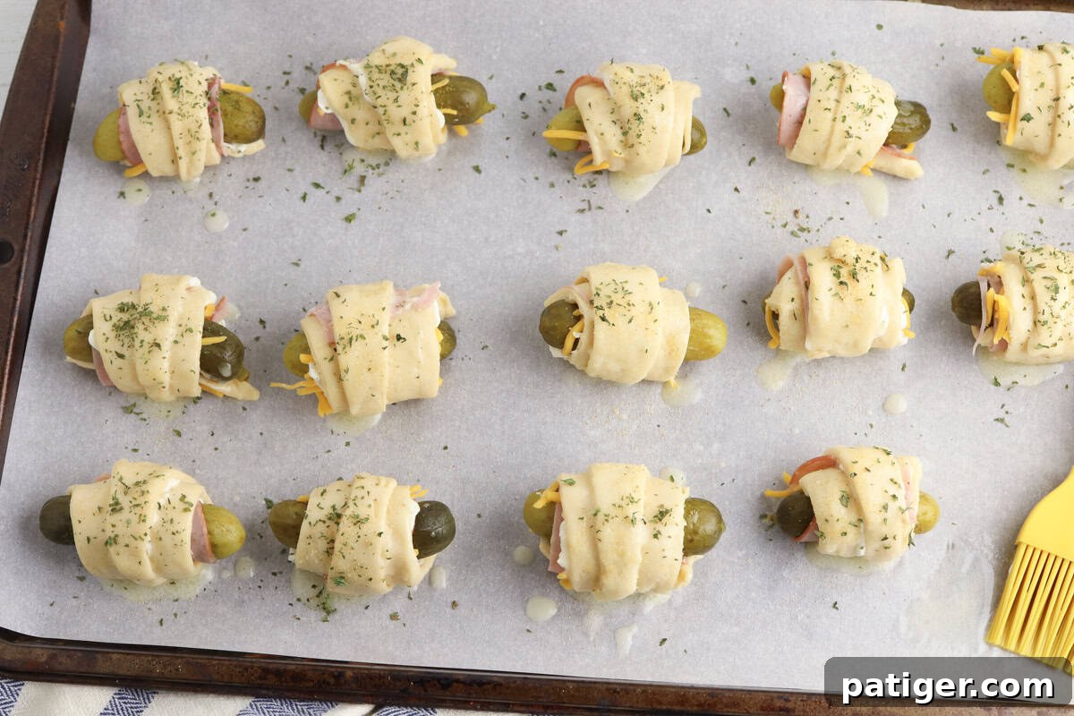 Dill Pickle Puffs 8 Pickles wrapped in crescent roll dough are evenly spaced on a parchment-lined baking sheet. The dough is topped with melted butter and sprinkled with herbs.