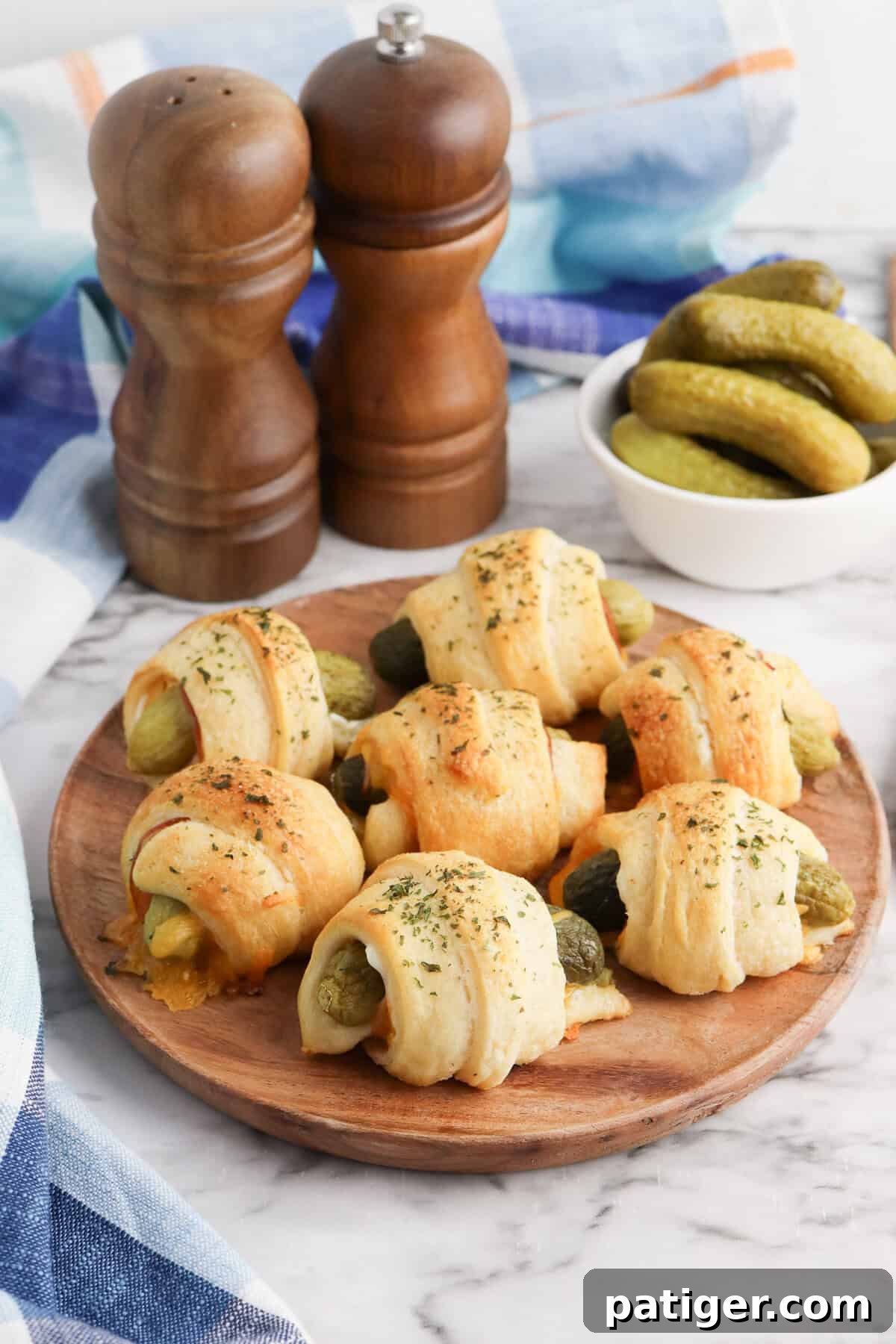 Dill Pickle Puffs 9 Pickles in a blanket on a wooden plate with melted cheese visible and bowl of mini dill pickles in the background.