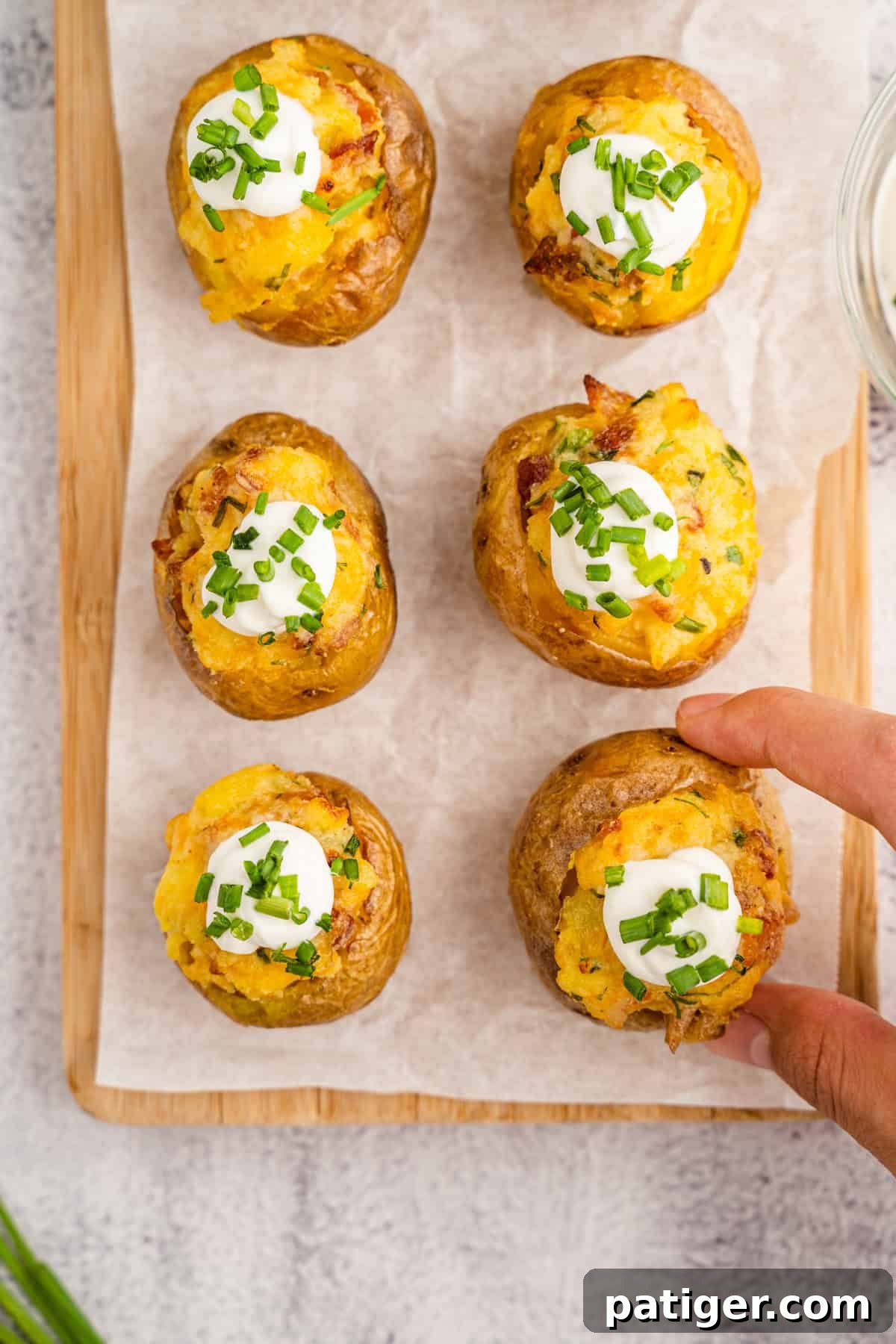 Loaded Potato Bites 10 Overhead view of six perfectly prepared twice-baked mini potatoes arranged on parchment paper, each generously garnished with creamy sour cream and vibrant fresh chopped chives. A hand reaches in, ready to grab one, highlighting their irresistible appeal.