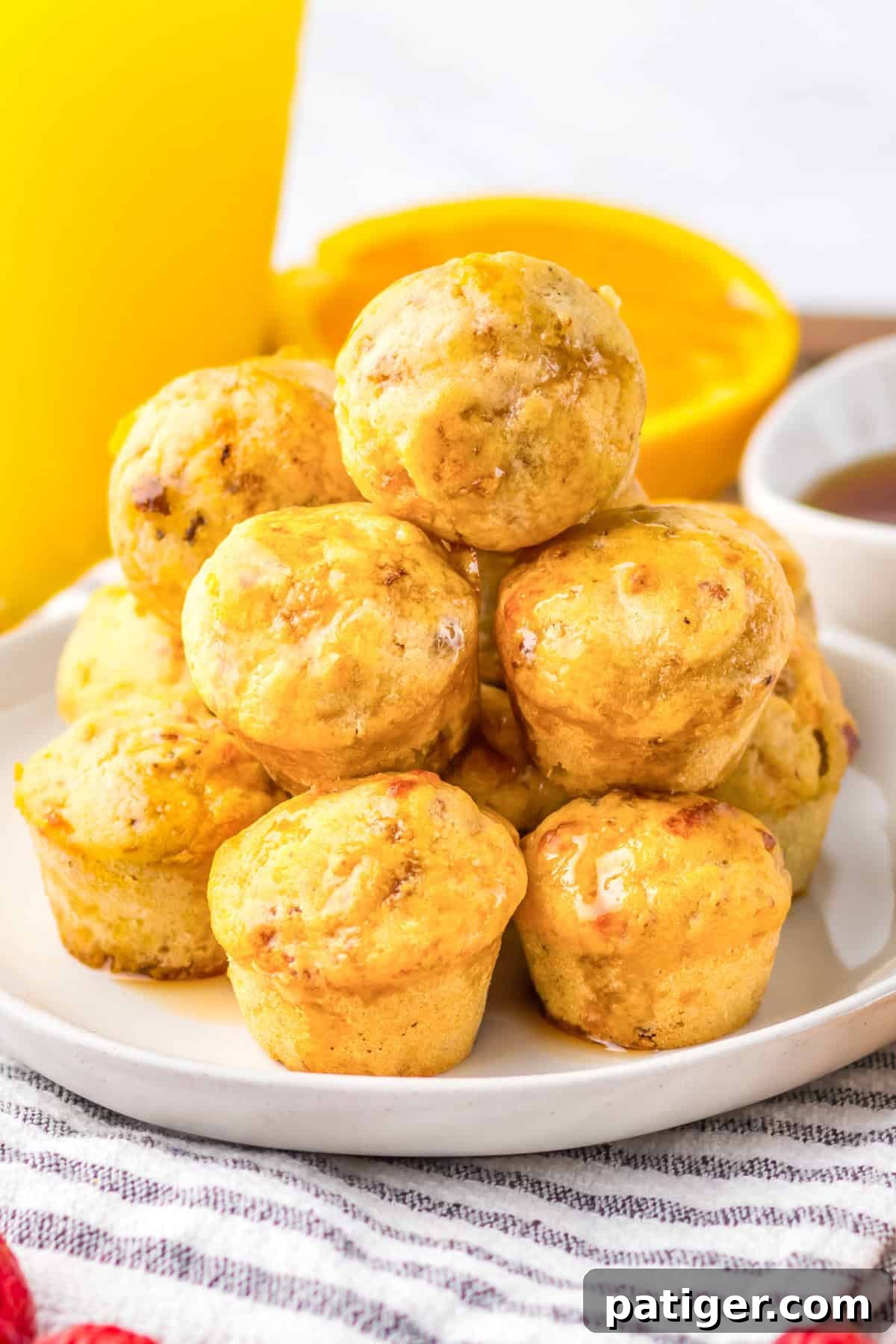 Mini sausage pancake muffins stacked on a plate, glistening with syrup. A glass of orange juice and syrup dish are blurred in the background.