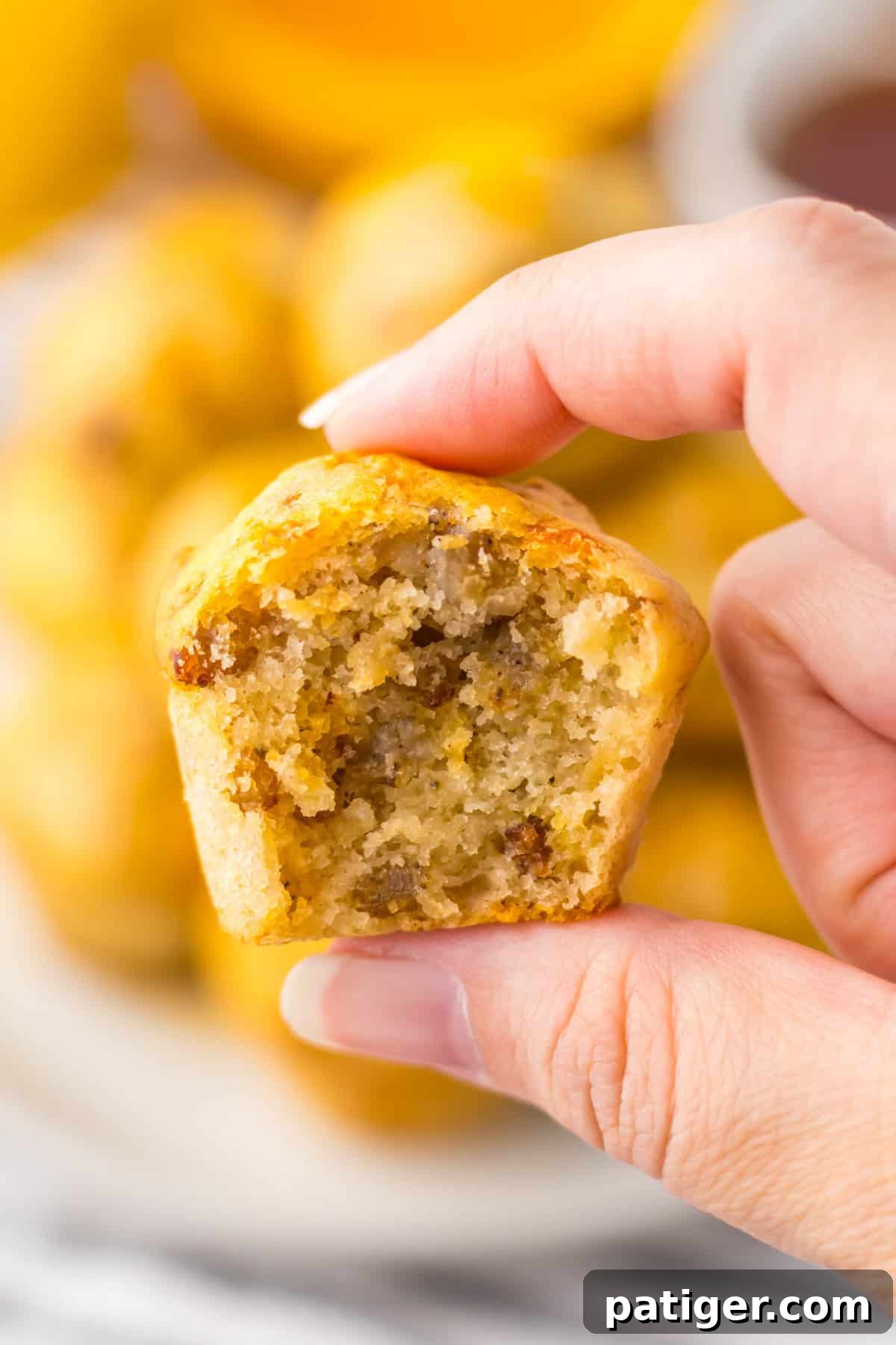 A hand holding a sausage McGriddle bite with a bite taken out, revealing a fluffy interior with pieces of sausage. The background shows more sausage pancake muffins stacked on a plate.