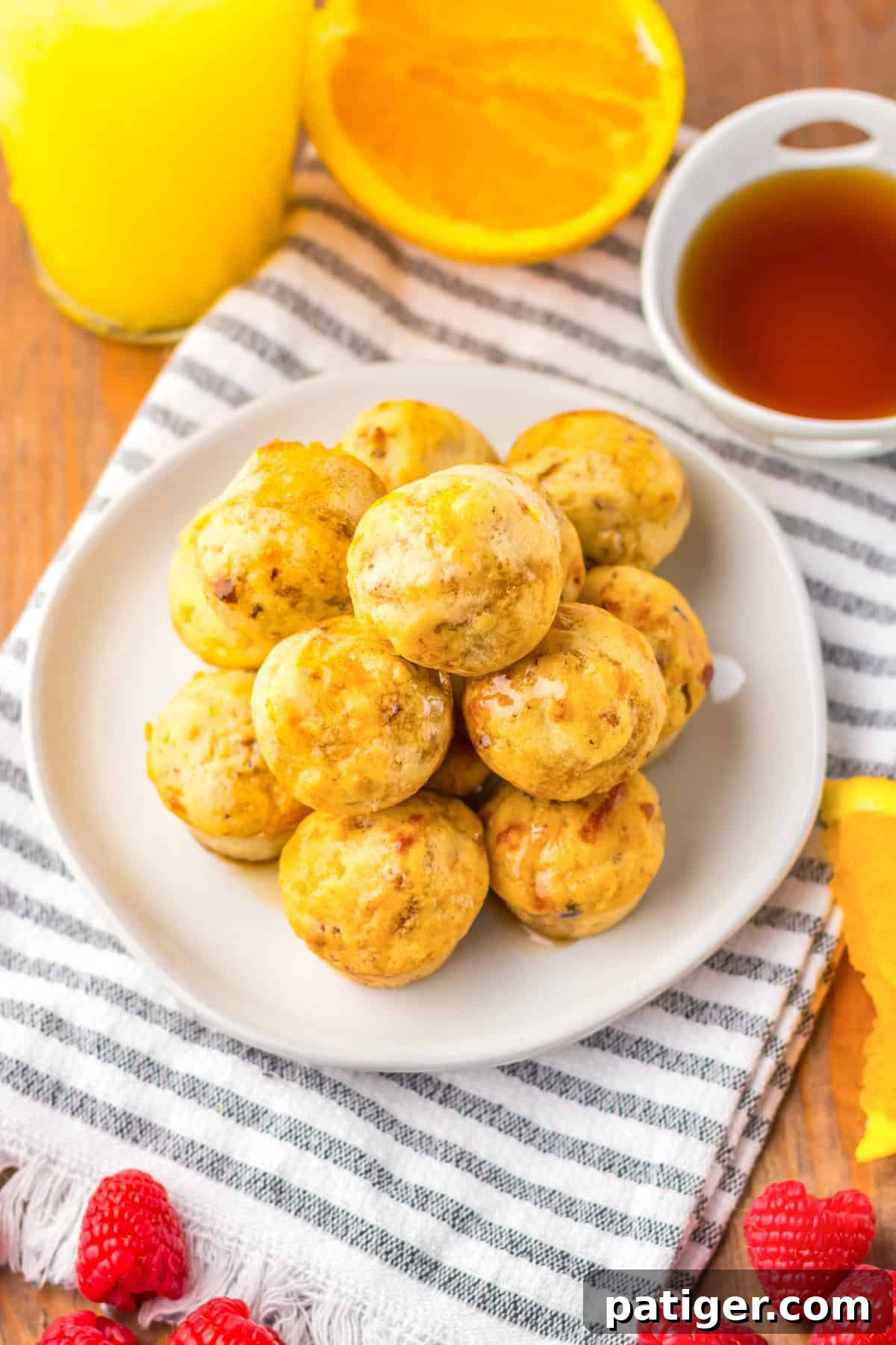 A plate of golden-brown sausage pancake muffins stacked high, drizzled with syrup. The plate sits on a striped cloth napkin with fresh raspberries, orange slices, a cup of syrup, and a glass of orange juice in the background.