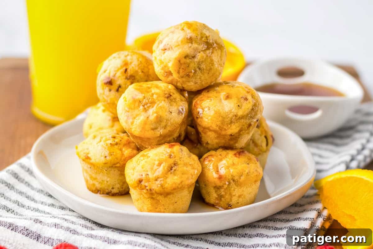 A stack of mini sausage and pancake muffins arranged on a white plate, drizzled with syrup. A glass of orange juice, a cup of syrup, and orange slices are in the background.