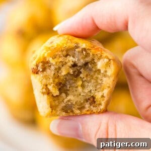A hand holding a mini sausage pancake muffin with a bite taken out, revealing a fluffy interior with pieces of sausage. The background shows more sausage McGriddle bites stacked on a plate.