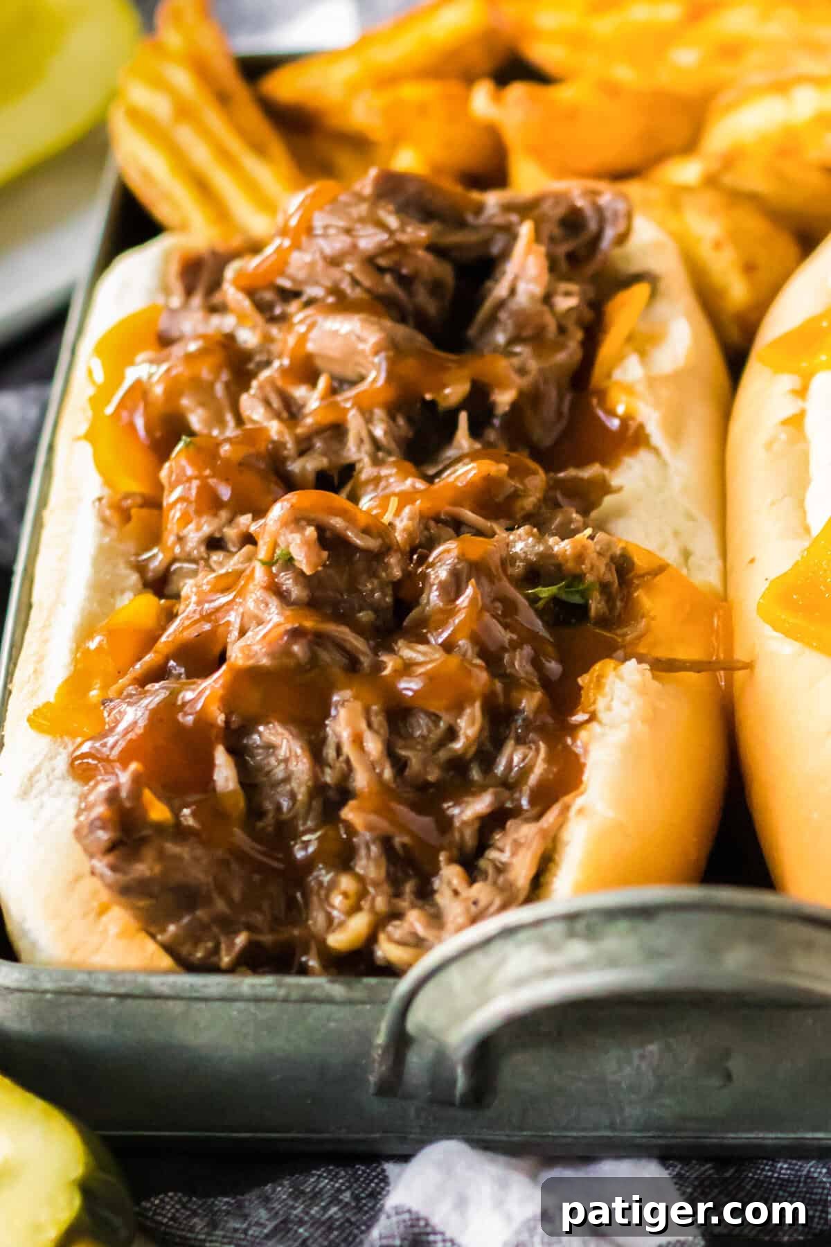 Melt-in-Your-Mouth Slow Cooker BBQ Beef 2 Tight close-up of one hoagie-style sandwich filled with shredded BBQ beef, cheddar cheese, and BBQ sauce, surrounded by fries and pickle slices.