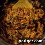 Melt-in-Your-Mouth Slow Cooker BBQ Beef 11 Close-up of shredded BBQ beef in a slow cooker, mixed with sauce and garnished with chopped green herbs. A wooden spoon is partially visible at the top.