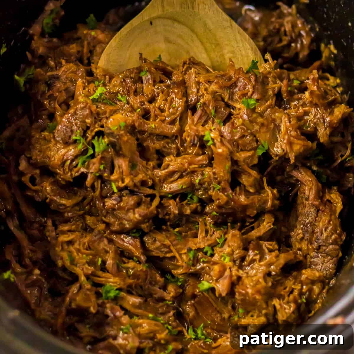 Melt-in-Your-Mouth Slow Cooker BBQ Beef 3 Close-up of shredded BBQ beef in a slow cooker, mixed with sauce and garnished with chopped green herbs. A wooden spoon is partially visible at the top.