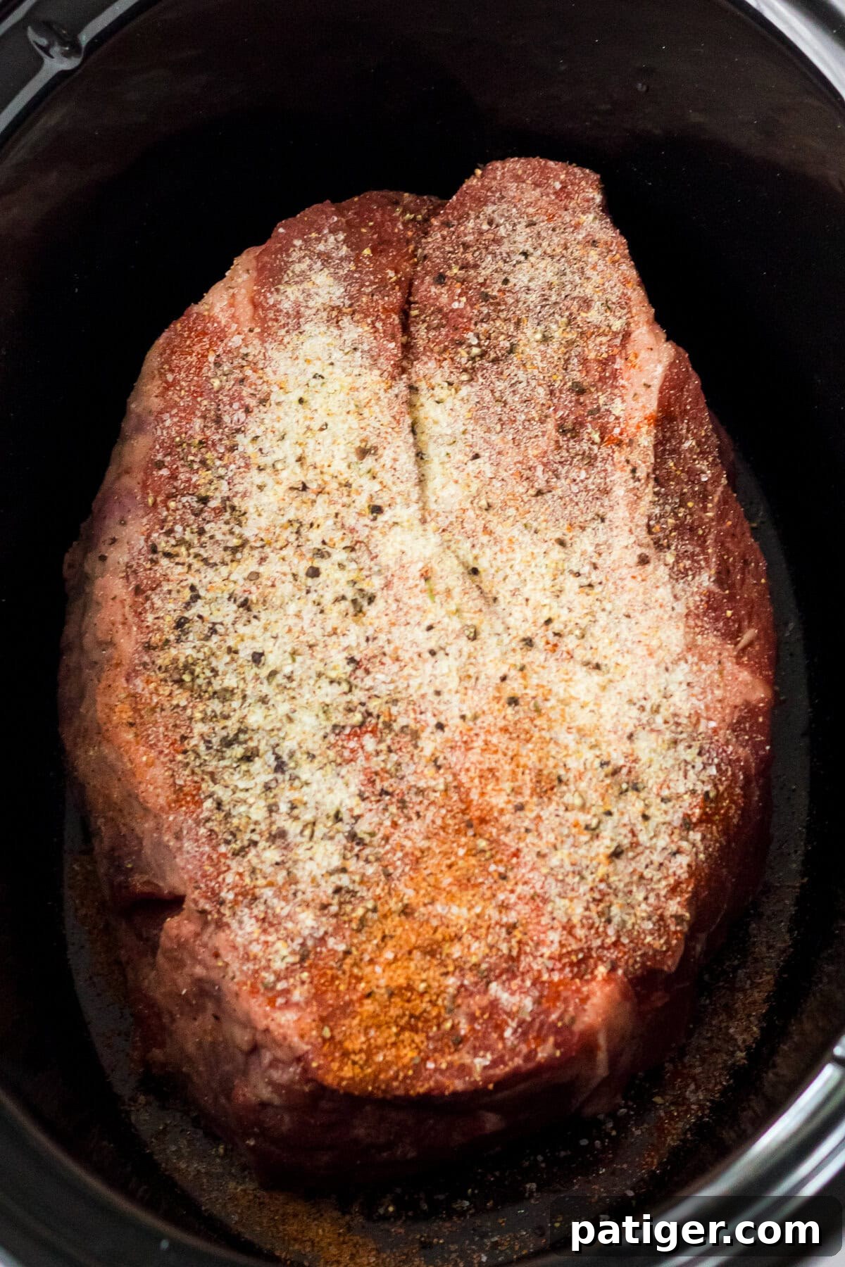 Melt-in-Your-Mouth Slow Cooker BBQ Beef 5 Overhead image of a large, raw chuck roast placed in a slow cooker, heavily seasoned with a dry rub of various spices including black pepper, paprika, and garlic powder.