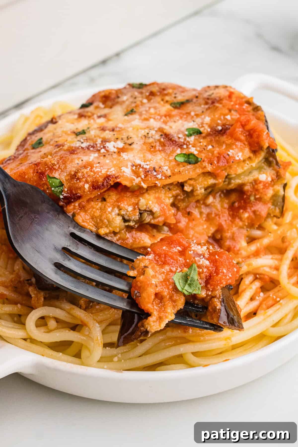 A close-up of a serving of baked eggplant Parmesan on a bed of spaghetti. A fork is lifting a bite, showing the saucy, cheesy, and layered eggplant with visible herbs and melted cheese on top.