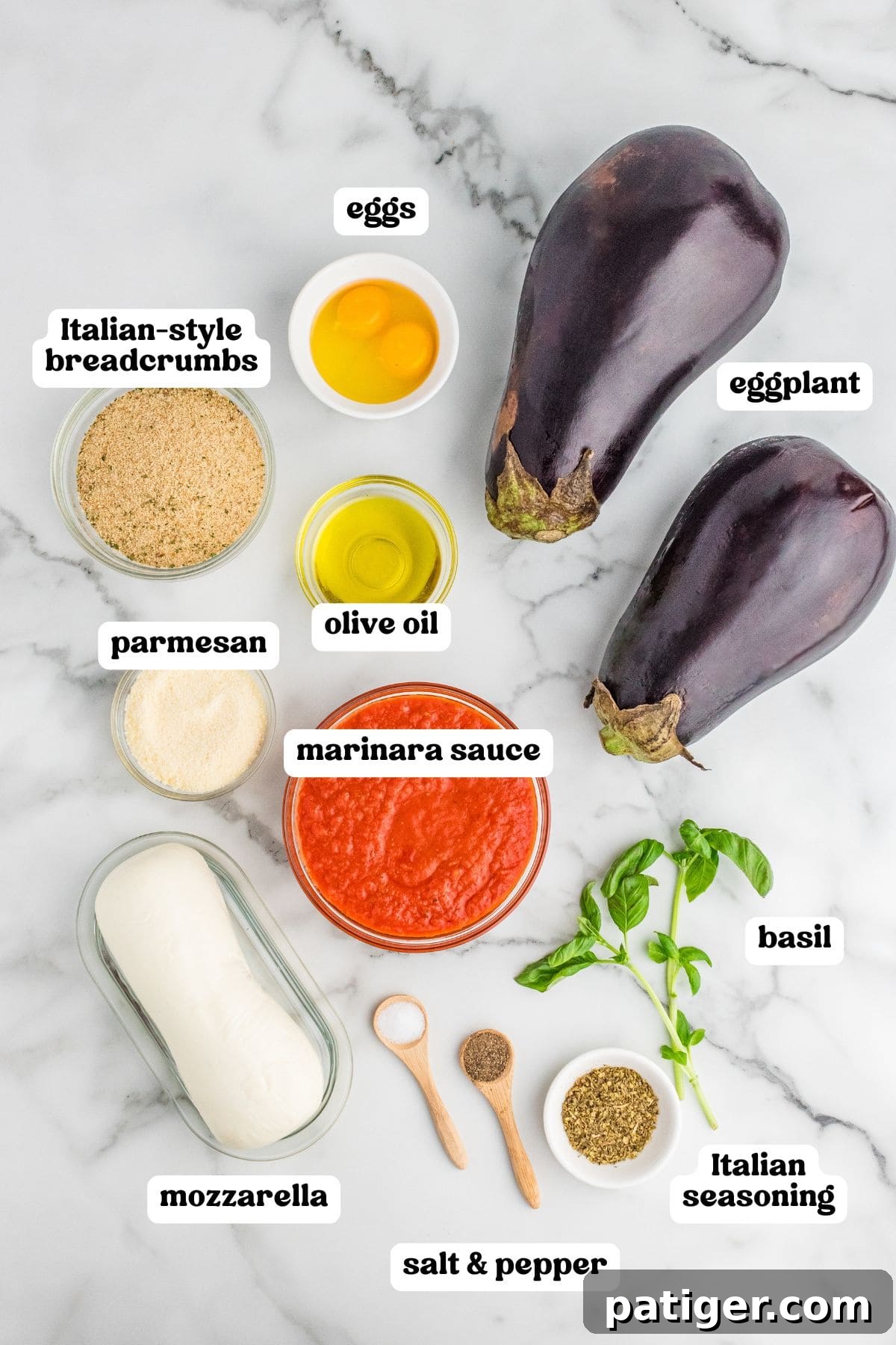 Overhead view of labeled ingredients for eggplant Parmesan: two whole eggplants, a bowl of marinara sauce, fresh mozzarella, grated parmesan, Italian-style breadcrumbs, olive oil, two eggs in a small bowl, a few sprigs of fresh basil, a small bowl of Italian seasoning, and wooden spoons holding salt and pepper.