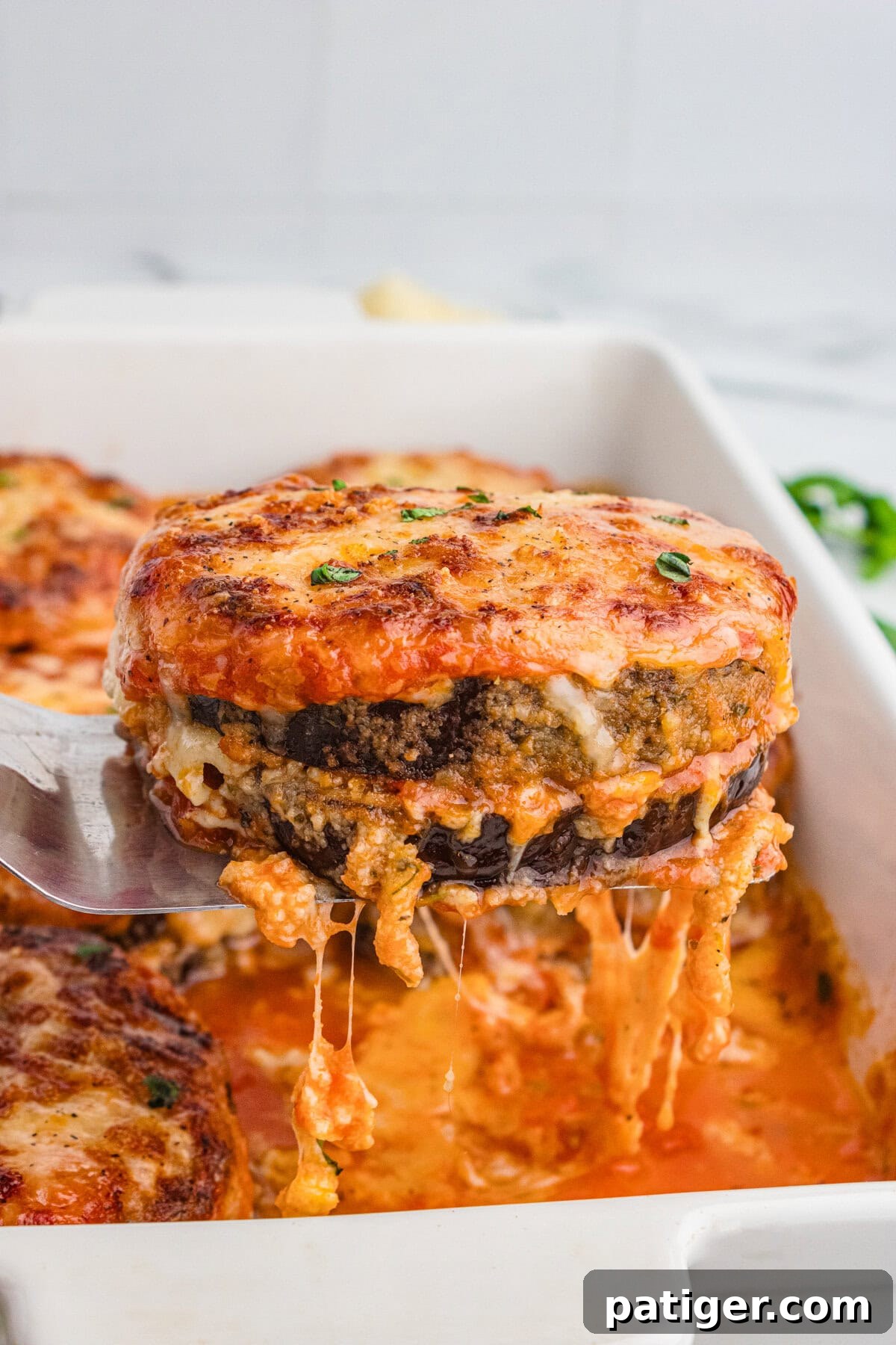 A cheesy slice of oven baked eggplant Parmesan being lifted from the baking dish, with stretchy cheese and visible layers of breaded eggplant, sauce, and cheese, garnished with fresh herbs.