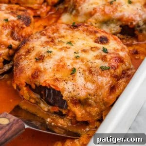 Close-up of a cheesy, golden-brown slice of baked eggplant Parmesan being lifted from a dish, revealing layers of eggplant and sauce with melted cheese on top, garnished with herbs.
