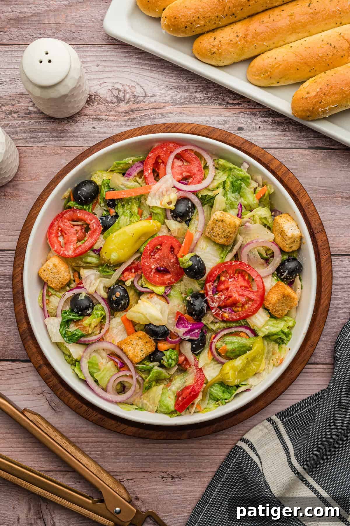 A beautifully arranged bowl of copycat Olive Garden house salad sits on a rustic wooden table. It features crisp lettuce, vibrant red tomatoes, thinly sliced red onions, glossy black olives, a few zesty pepperoncini, crunchy croutons, and a generous sprinkle of shredded Parmesan cheese, perfectly dressed with Italian vinaigrette. Fresh breadsticks are visible on a white platter in the background, along with salt and pepper shakers.
