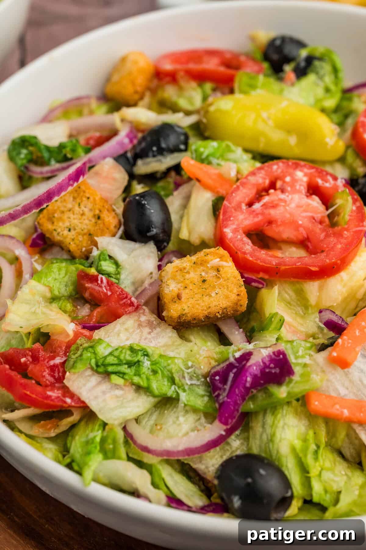 A close-up view of the copycat Olive Garden Salad, highlighting the intricate details of each ingredient. Crunchy croutons, juicy red tomato slices, pitted black olives, thin rings of red onion, and vibrant green pepperoncini are all coated in a rich, creamy Italian dressing, making it almost indistinguishable from the restaurant's original.