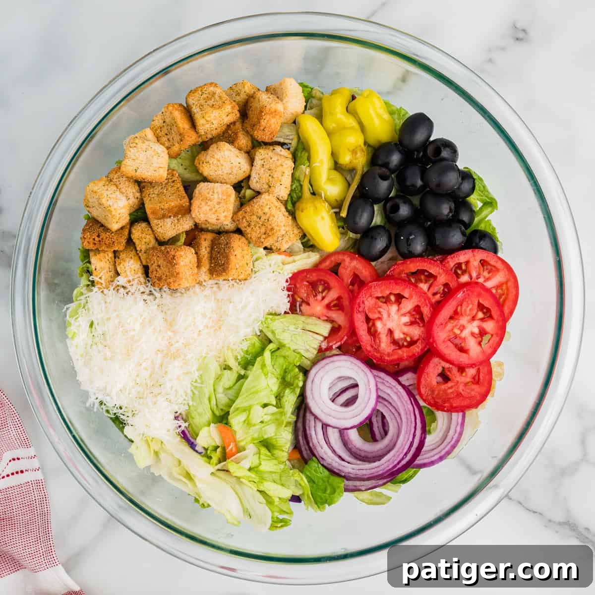 A large clear glass bowl filled with the base ingredients of the Olive Garden copycat salad. Crisp iceberg lettuce forms the bottom layer, topped with a colorful assortment of sliced Roma tomatoes, whole pitted black olives, delicate rings of red onion, vibrant green pepperoncini, and a generous scattering of golden croutons and freshly shredded Parmesan cheese. All components are neatly arranged and ready for the dressing.