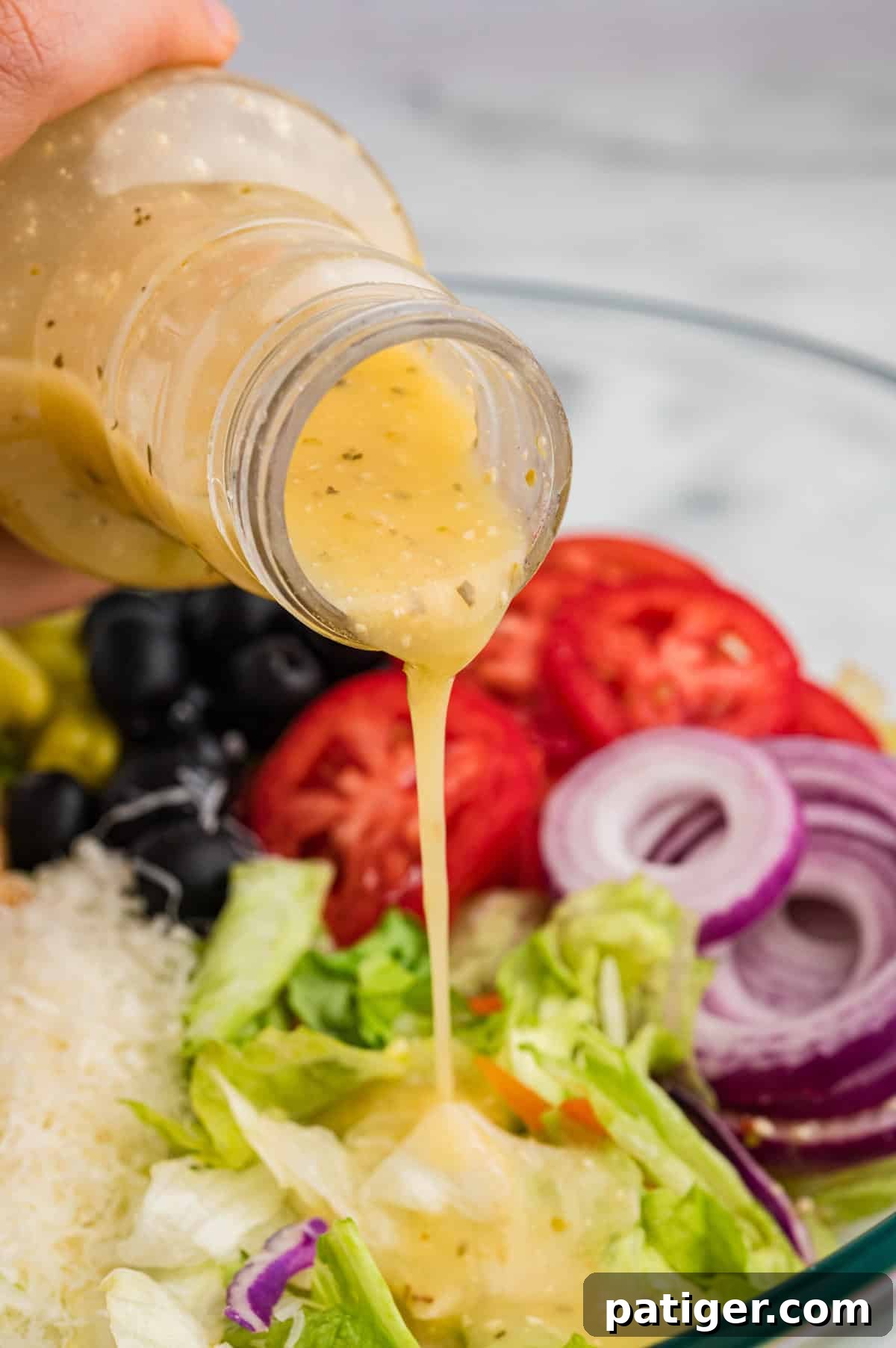 A close-up shot capturing the moment Olive Garden signature Italian dressing is being poured from its distinctive bottle onto a vibrant, fresh salad. The salad features crisp iceberg lettuce, perfectly sliced red tomatoes, delicate rings of red onion, shiny pitted black olives, bright green pepperoncini, and a dusting of grated Parmesan cheese, all eagerly awaiting the flavorful dressing to bind them together.