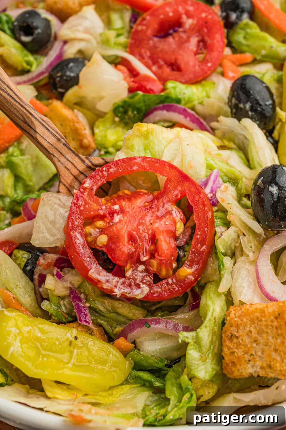 A close-up shot of a perfectly tossed Olive Garden Italian Salad, showcasing a vibrant cross-section of ingredients. A juicy slice of tomato rests on a bed of crisp iceberg lettuce, surrounded by glossy black olives, delicate red onion rings, golden-brown croutons, finely shredded carrots, and a bright yellow pepperoncini. The elements are beautifully coated in dressing, ready to be enjoyed.