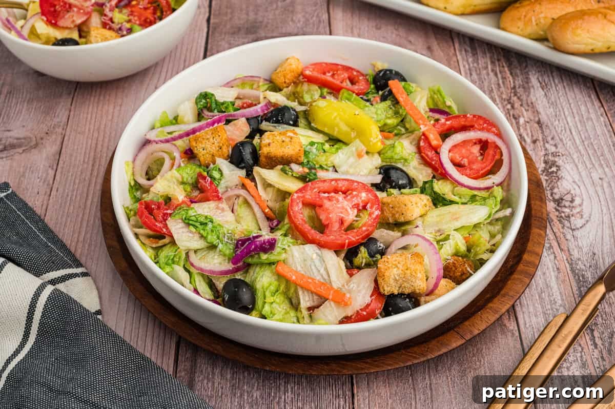 A generously sized bowl of copycat Olive Garden salad rests on a rustic wooden table, adorned with a black-and-white striped napkin. The salad is vibrant and inviting, packed with crisp lettuce, juicy sliced tomatoes, pitted black olives, thin rings of red onion, zesty pepperoncini, crunchy croutons, and a sprinkle of shredded cheese, all gleaming with dressing. In the soft-focus background, a plate of garlic breadsticks and salad tongs complete the enticing Italian meal scene.
