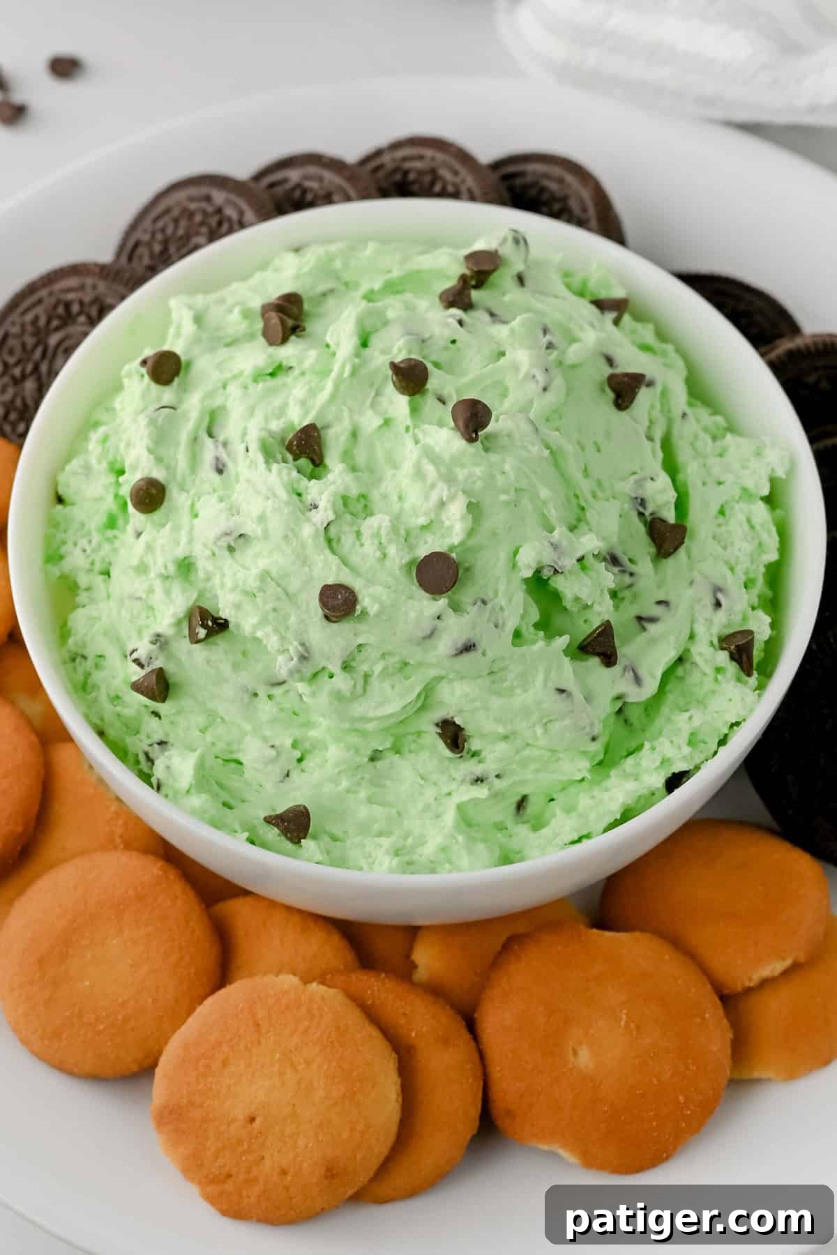 A bowl of creamy green mint chocolate chip dip topped with scattered chocolate chips, served with vanilla wafers and chocolate sandwich cookies for dipping.