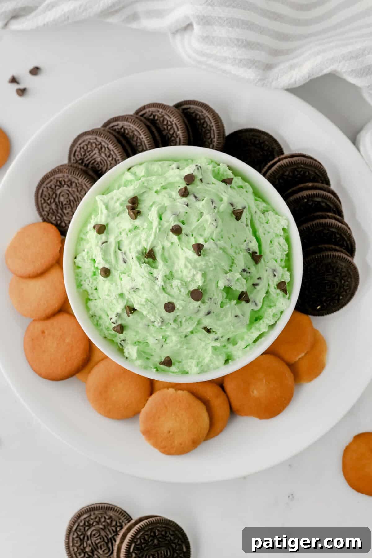  Top-down view of a white bowl filled with mint chocolate chip dip with chocolate chips on top, placed on a white plate with a ring of chocolate sandwich cookies on one side and vanilla wafers on the other.