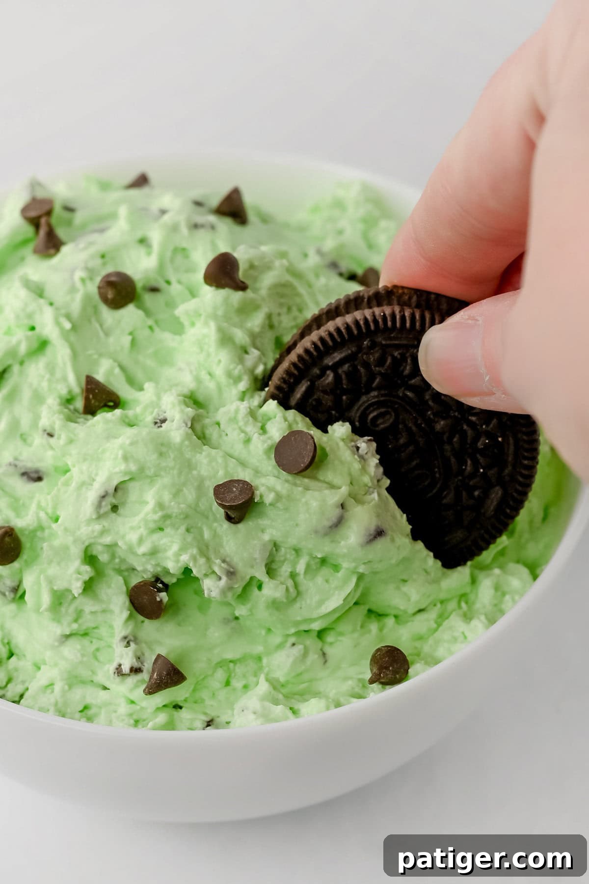 A hand holding an OREO cookie, dipping it into a bowl of green mint chocolate chip dip with chocolate chips.