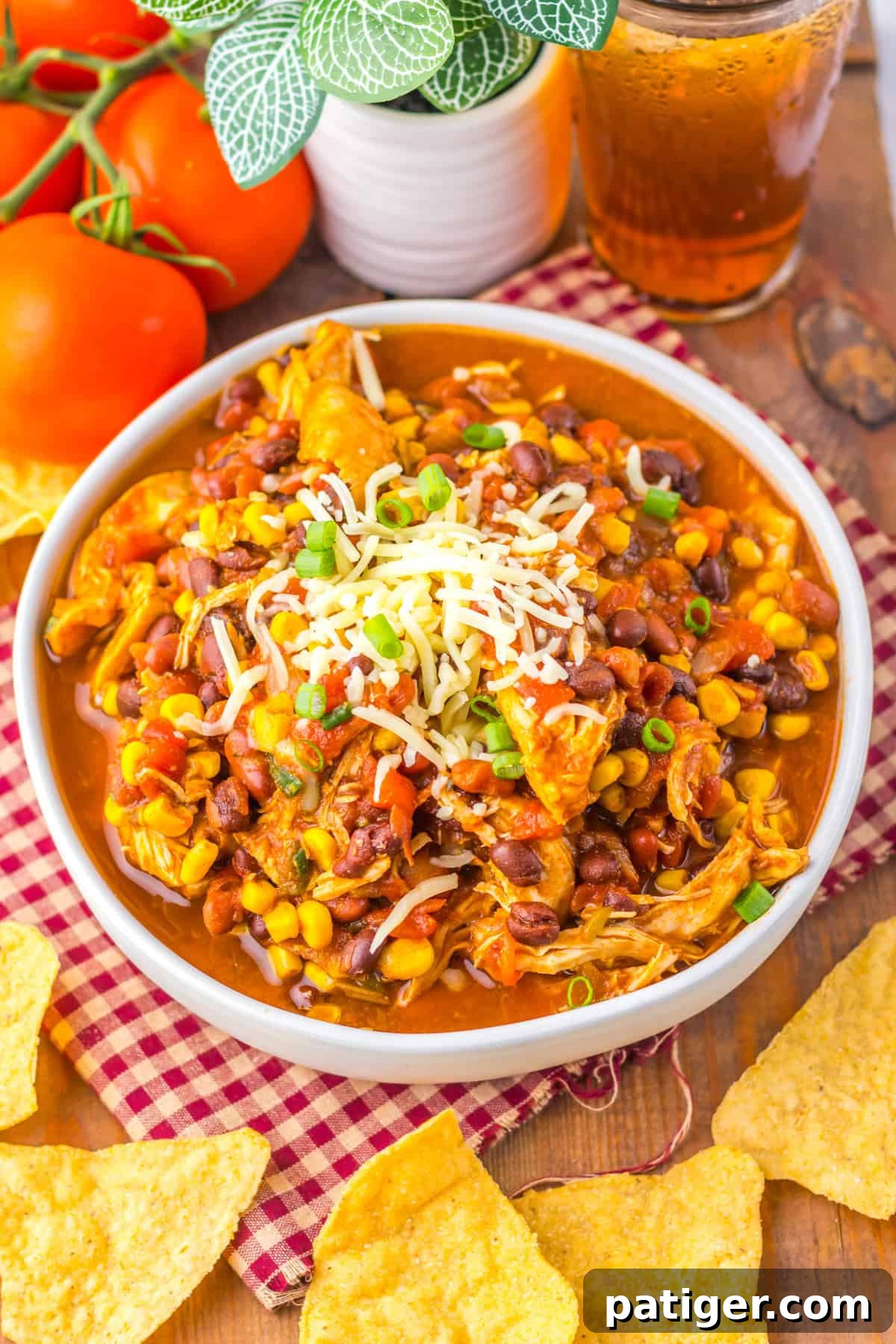 A bowl of slow cooker chicken taco soup, featuring shredded chicken, corn, beans, diced tomatoes, and topped with shredded cheese and green onions. The bowl is placed on a red and white checkered cloth, surrounded by tortilla chips, fresh tomatoes, and a glass of iced tea.