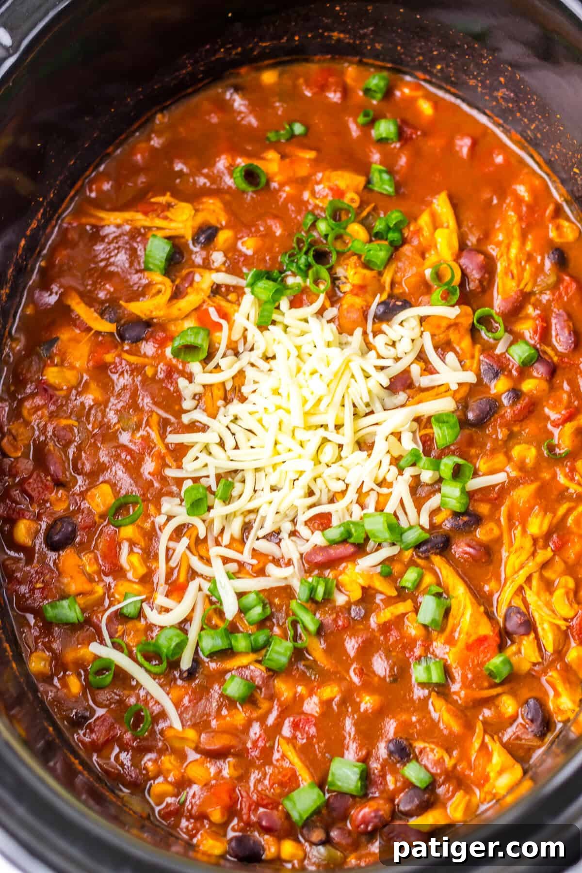  An overhead view of slow cooker taco soup with chicken inside a black crockpot. The soup is topped with shredded cheese and chopped green onions, featuring visible ingredients like shredded chicken, black beans, corn, and diced tomatoes in a hearty broth.