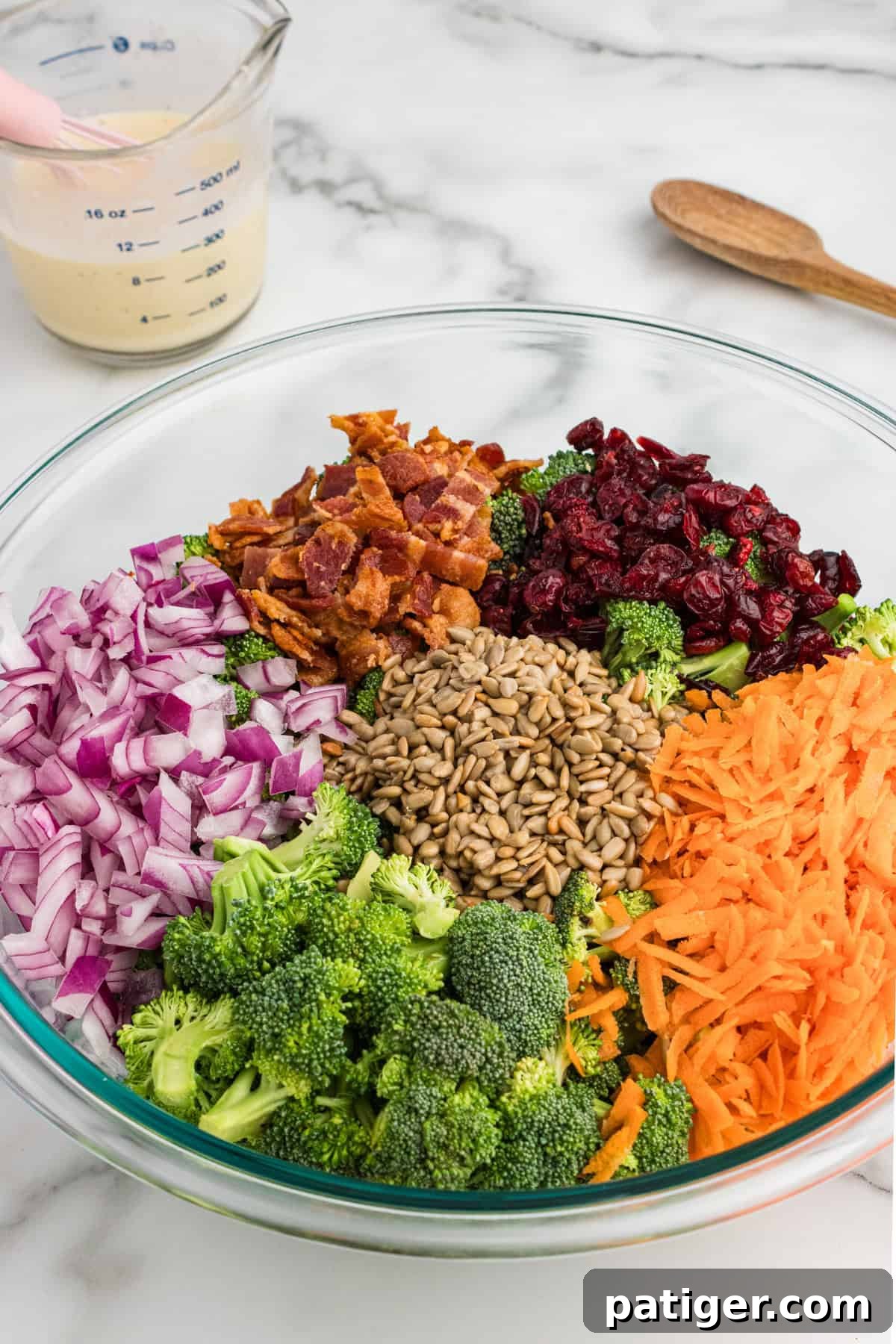A large glass bowl containing all the dry ingredients for broccoli crunch salad: vibrant green broccoli florets, shredded orange carrots, diced red onions, crispy bacon pieces, plump dried cranberries, and golden sunflower seeds, all mixed together.
