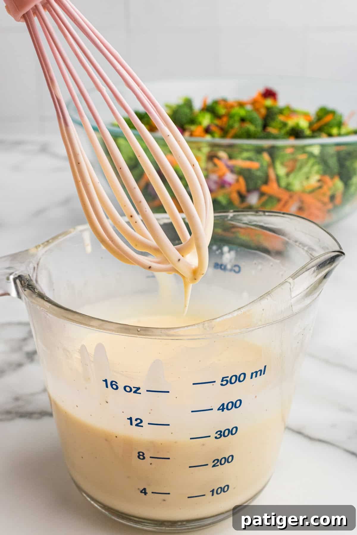 A glass measuring cup holding a generous amount of creamy, pale yellow dressing with a whisk resting inside, indicating it has just been mixed.