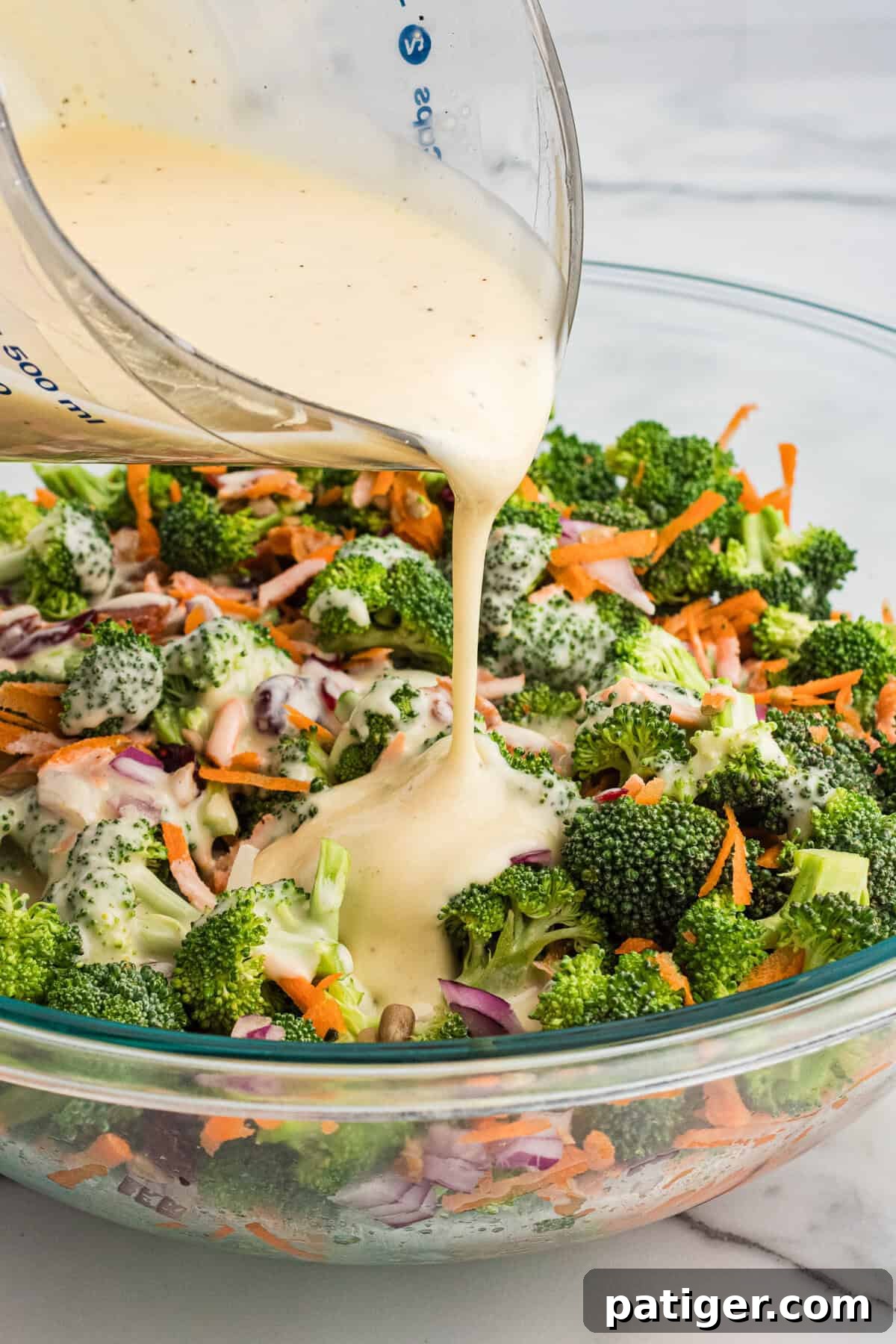 A close-up shot of the creamy dressing being poured from a clear glass measuring cup over a colorful bowl of broccoli crunch salad, showing the rich coating forming on the vegetables.