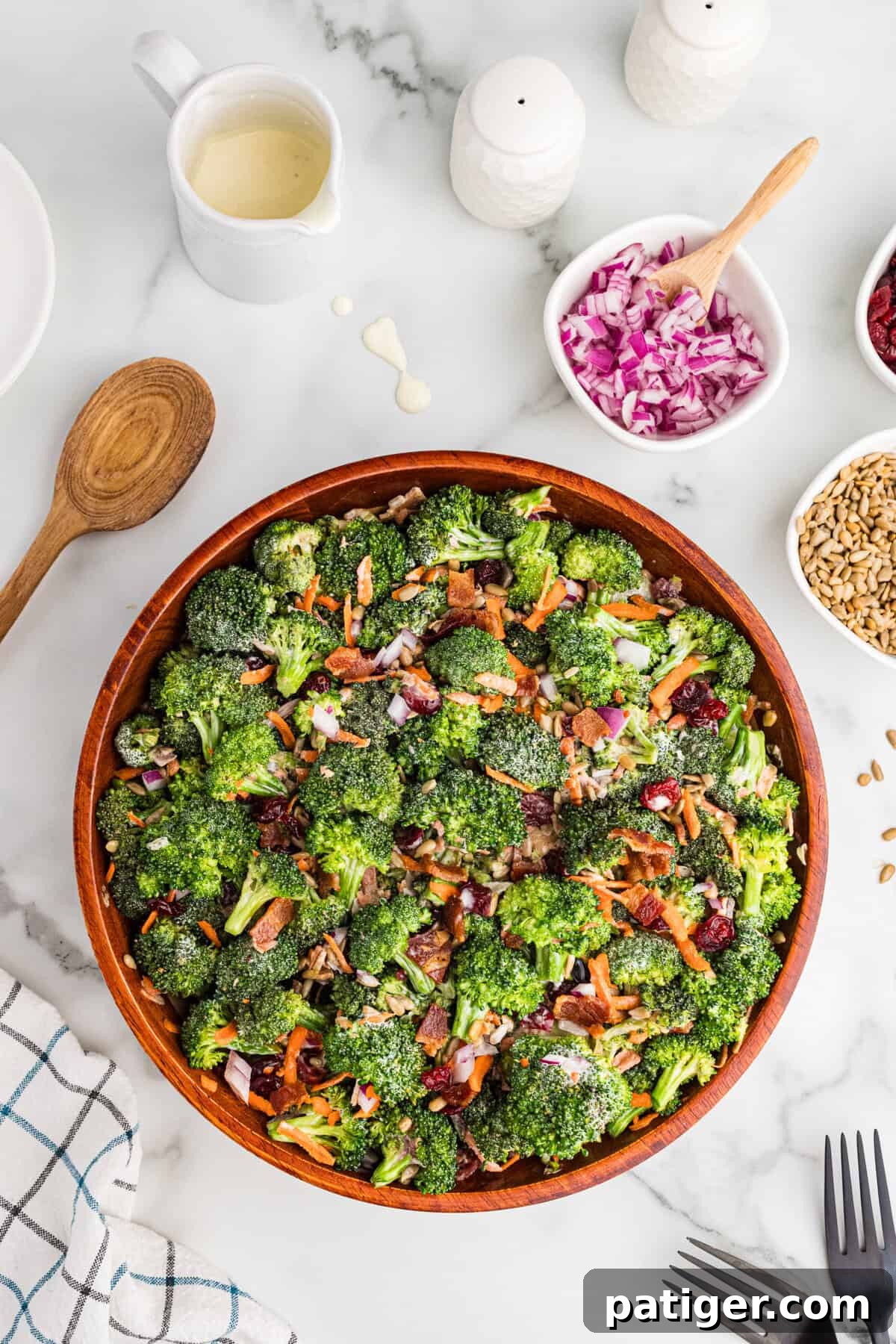 A top-down, beautifully styled view of a large rustic wooden bowl brimming with vibrant broccoli crunch salad. The salad is composed of fresh broccoli, crispy bacon, ruby red cranberries, finely shredded carrots, golden sunflower seeds, and thinly sliced red onions. Surrounding the bowl are small, elegant dishes holding individual extra ingredients, a rustic wooden spoon, and a charming checkered kitchen towel, all arranged on a pristine marble surface.
