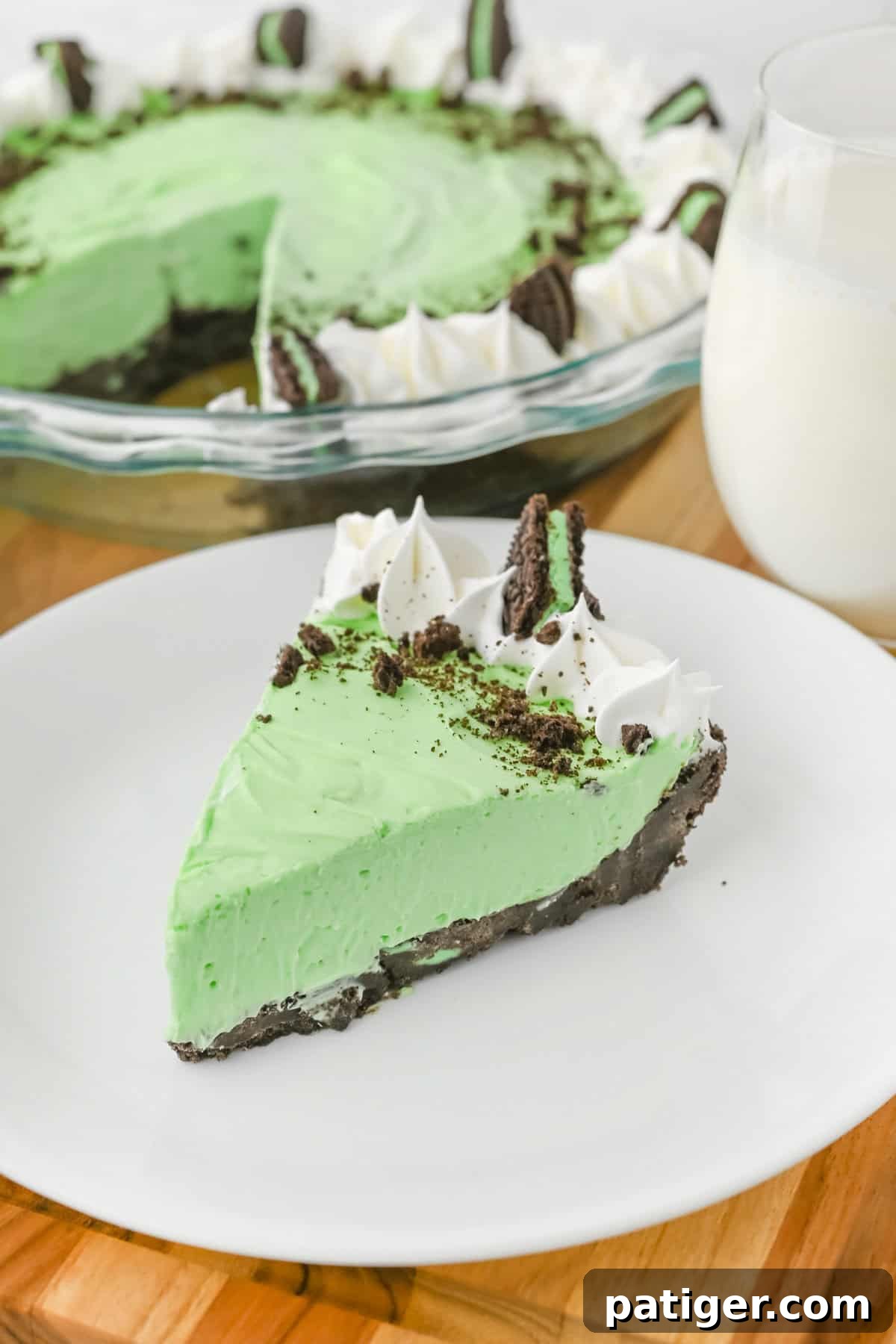 A perfectly sliced no-bake grasshopper pie, showcasing its smooth, vibrant green filling, robust chocolate cookie crust, and a lavish topping of whipped cream, crushed cookies, and a delicate Mint Oreo piece. The full pie and a glass of milk are softly blurred in the background, set on a pristine white plate.