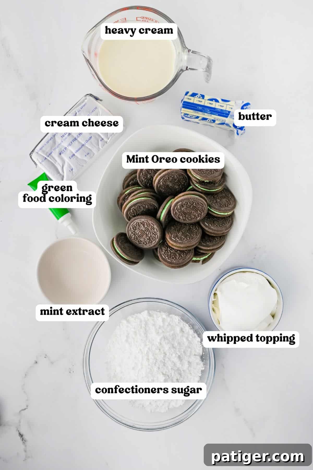 A top-down view displaying all the labeled ingredients needed for a Grasshopper Mint Pie, neatly arranged: heavy cream, softened cream cheese, melted butter, a small container of green food coloring, a stack of Mint Oreo cookies, a bottle of mint extract, a tub of whipped topping, and a bowl of confectioners sugar.