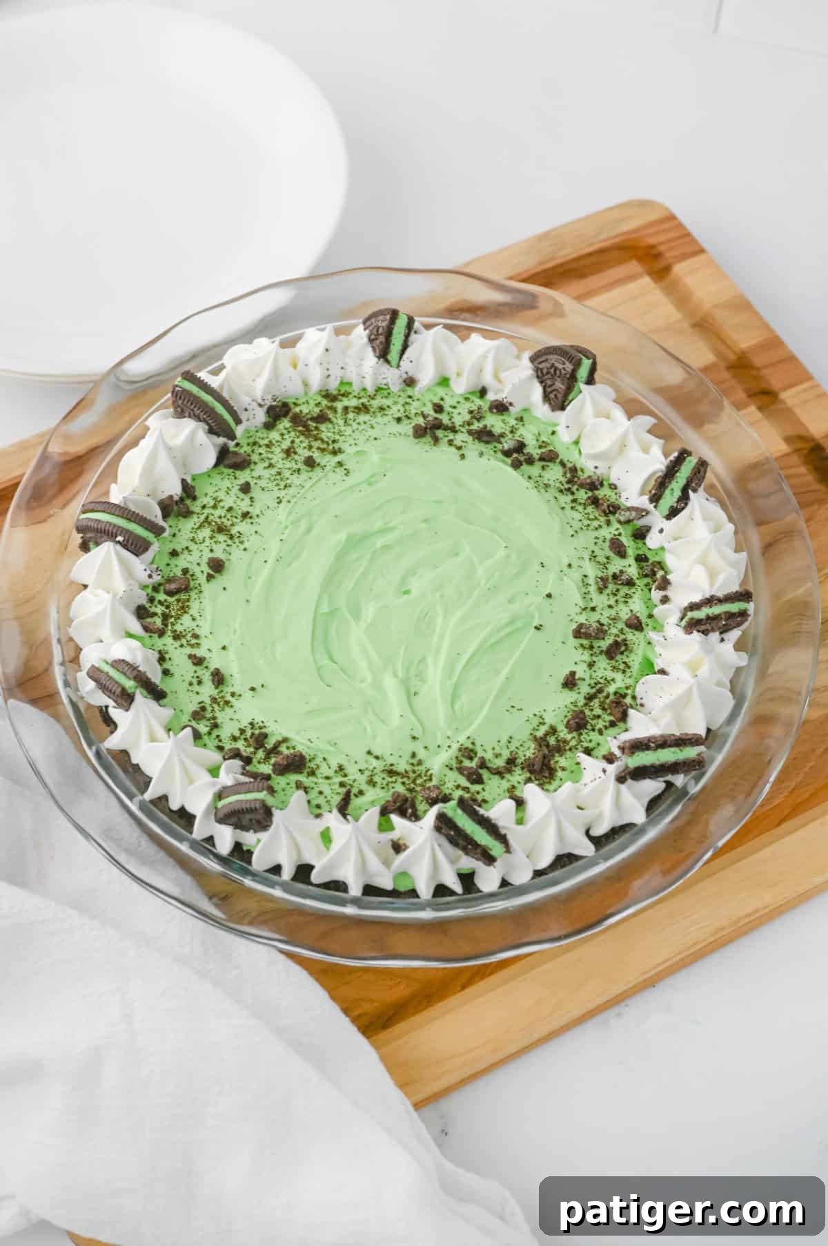 A complete No-Bake Grasshopper Mint Pie in a clear glass pie dish, generously topped with swirling whipped cream, a sprinkle of crushed chocolate cookies, and several halved Mint Oreo cookies for decoration. The green filling is visible beneath the toppings.
