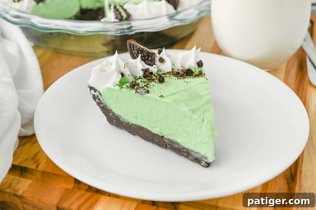 A beautifully presented frozen mint Grasshopper pie slice, featuring its vibrant green filling, rich chocolate crust, decorative swirls of whipped cream, a sprinkling of crushed cookies, and a whole Mint Oreo piece on top. It's served on a clean white plate, with the full pie in a glass dish and a glass of milk softly blurred in the background.