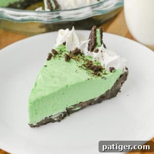 A close-up of a slice of no bake grasshopper pie with green filling, chocolate crust, whipped cream swirls, crushed chocolate cookies, and a Mint Oreo half, served on a white plate.