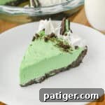 A close-up of a slice of no bake grasshopper pie with green filling, chocolate crust, whipped cream swirls, crushed chocolate cookies, and a Mint Oreo half, served on a white plate.