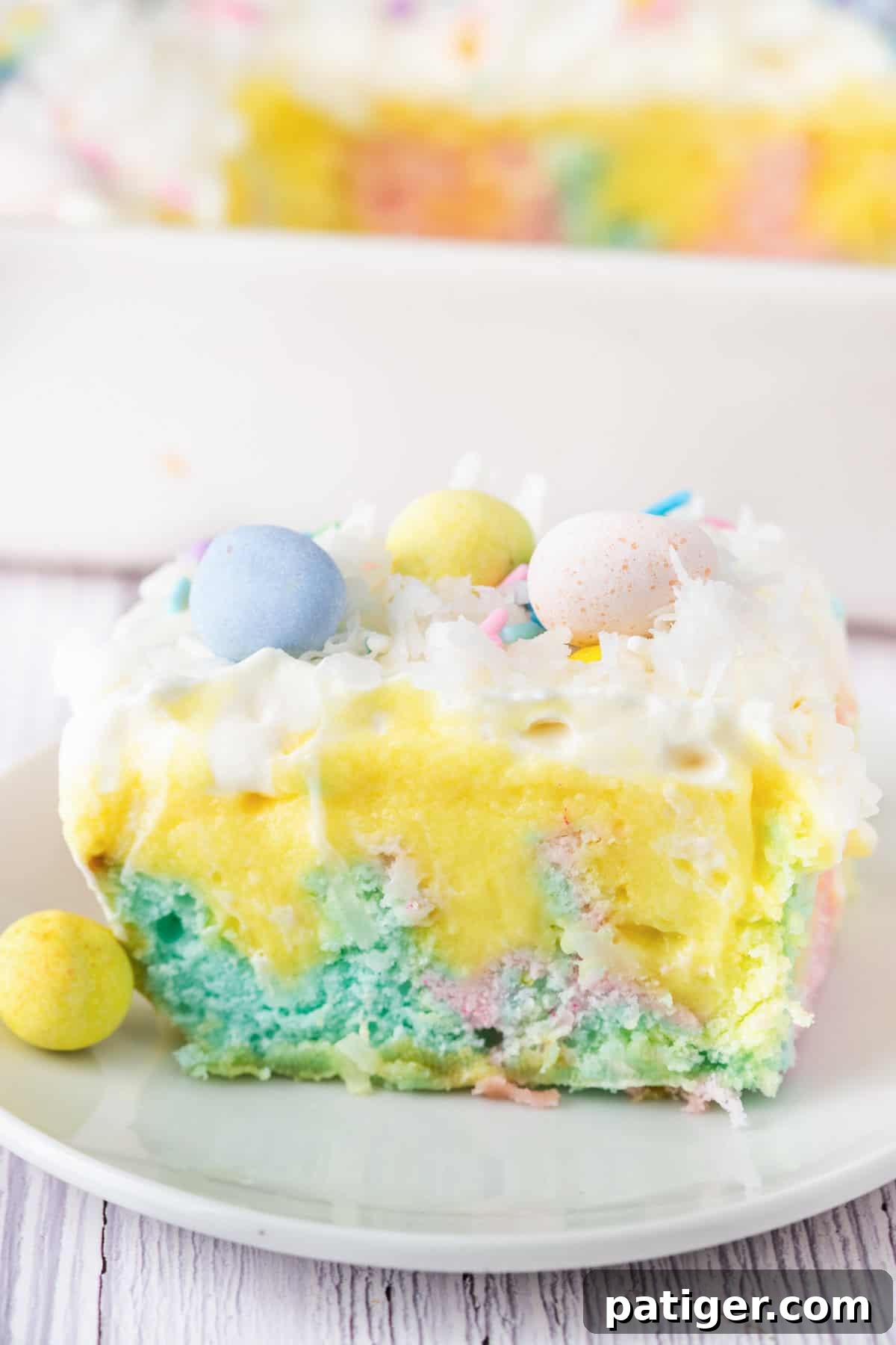 A pastel Easter poke cake slice on a plate, showing its colorful interior of pink, blue, and yellow cake, topped with pudding, whipped cream, coconut, and mini chocolate eggs. A blurred baking dish with more cake is in the background.