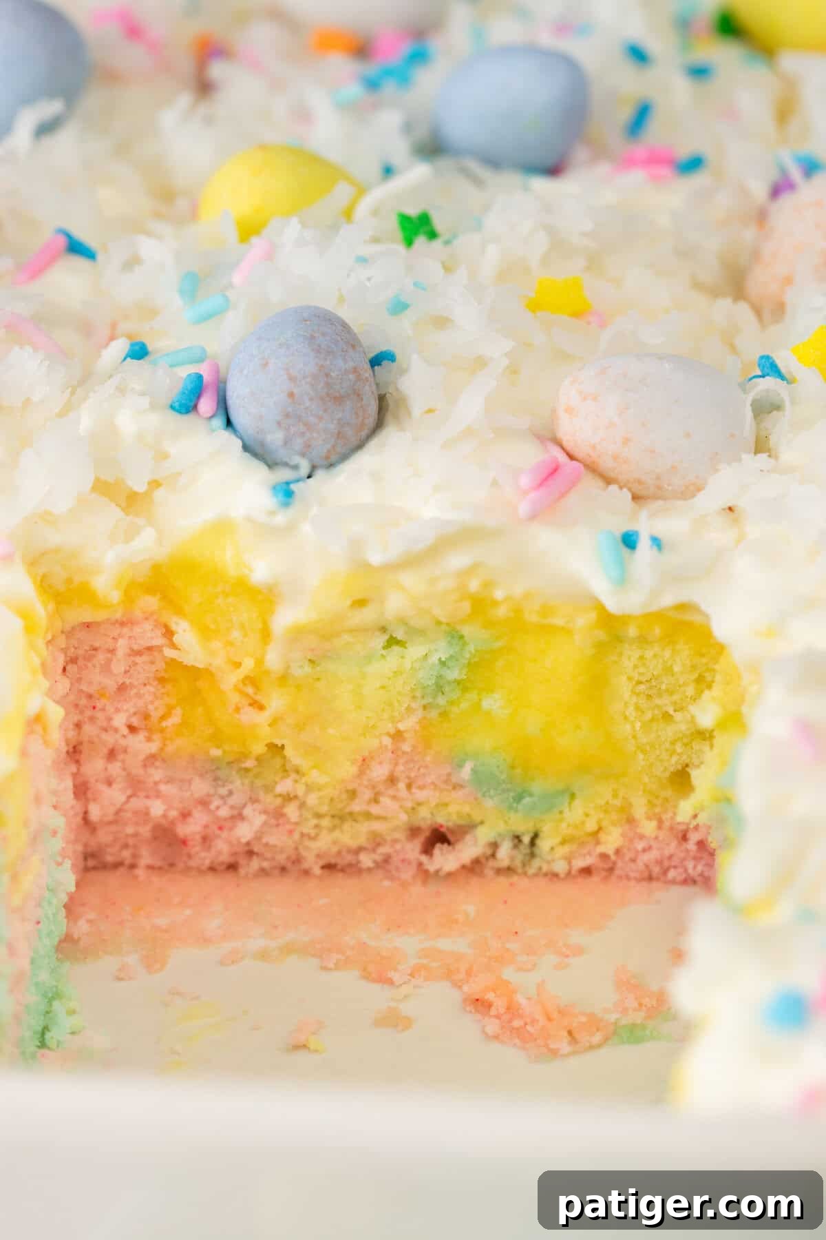 A close-up of a partially eaten Easter poke cake in a baking dish. The cake has pastel pink, yellow, and blue layers with yellow pudding filling and is topped with whipped cream, shredded coconut, pastel sprinkles, and mini chocolate eggs.