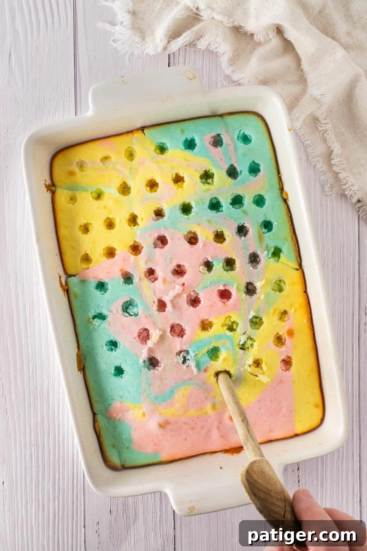 Baked pastel Easter cake in a white baking dish with holes poked throughout using a wooden spoon. The cake has swirled pink, blue, and yellow layers.baked pastel Easter cake in a white baking dish with holes poked throughout using a wooden spoon. The cake has swirled pink, blue, and yellow layers.