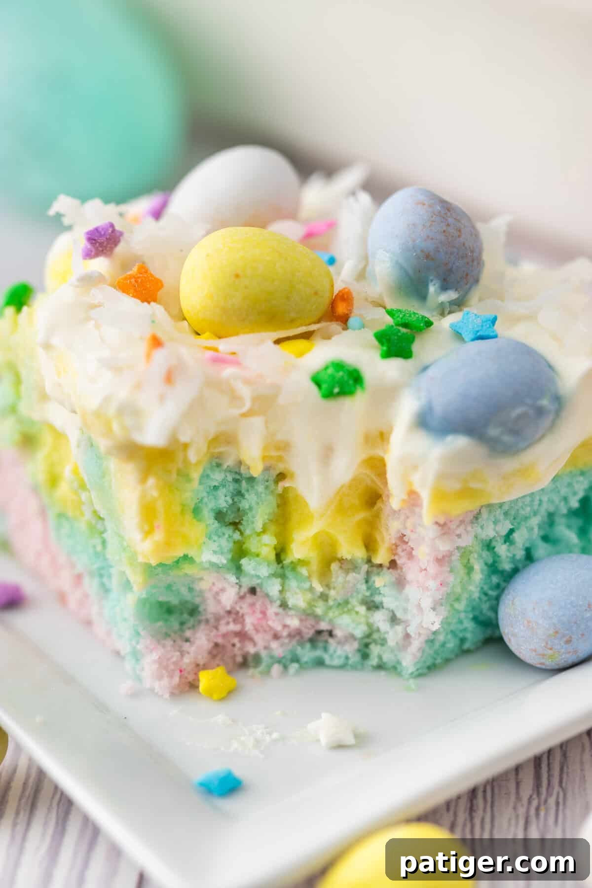 A slice of Easter poke cake with pudding on a white plate with a bite taken out. The cake has swirled pink, blue, and yellow layers, with vanilla pudding, whipped cream, coconut, colorful sprinkles, and mini chocolate eggs as toppings.