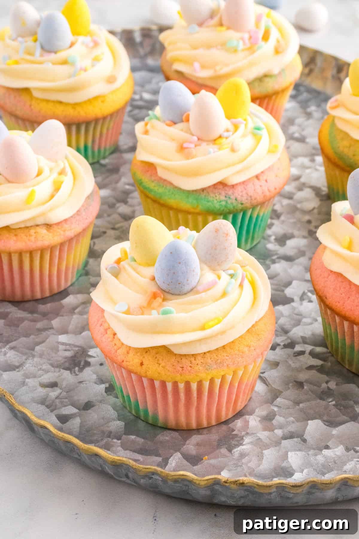 Hoppy Easter Cupcake Treats 2 Several pastel swirl Easter cupcakes sit on a round metal tray, each topped with piped buttercream frosting, pastel sprinkles, and mini chocolate eggs.