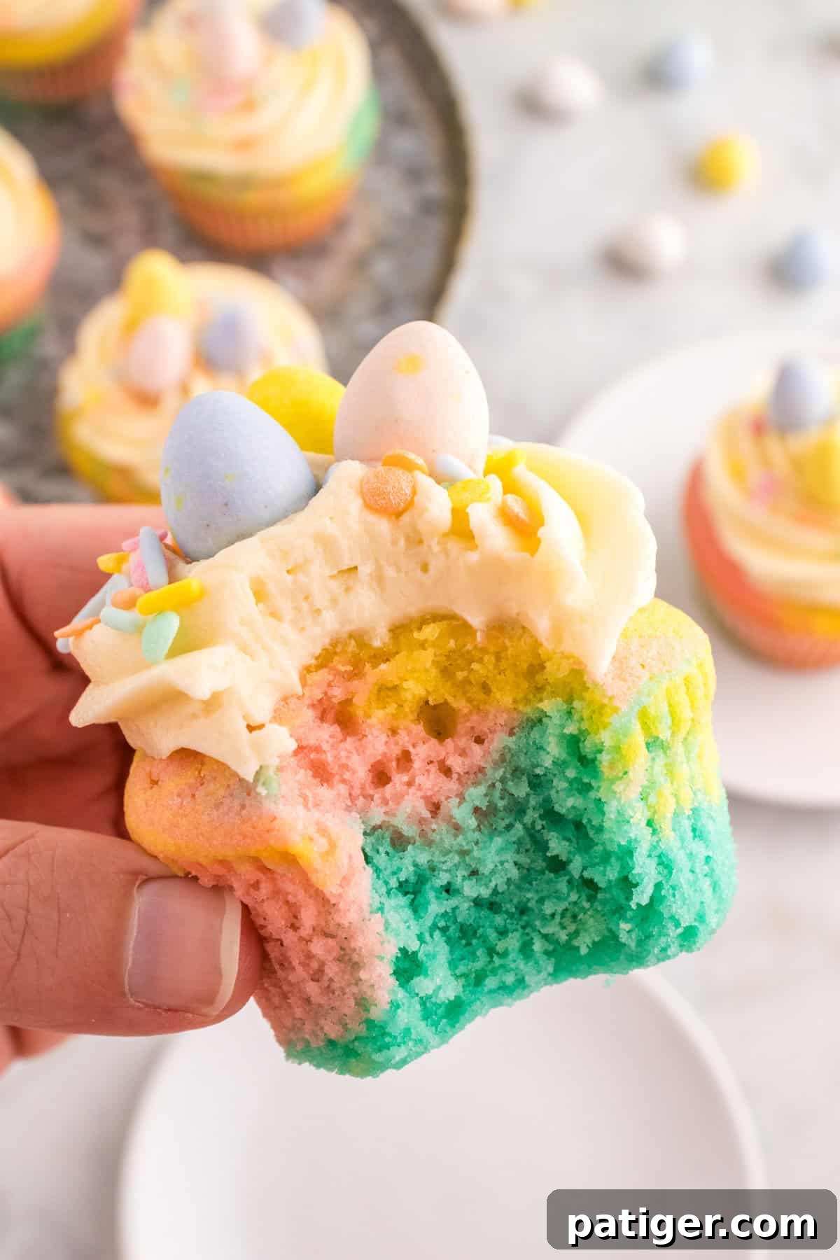 Hoppy Easter Cupcake Treats 11 A pastel swirl cupcake held in a hand, showing the colorful blue, pink, and yellow layers inside. The cupcake is decorated with buttercream frosting, pastel sprinkles, and three mini chocolate eggs.