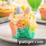 A close-up of a pastel swirl Easter cupcake with a bite taken out, revealing soft layers of blue, pink, and yellow cake. The cupcake is topped with piped buttercream frosting, pastel sprinkles, and three mini chocolate eggs in blue, yellow, and pink.